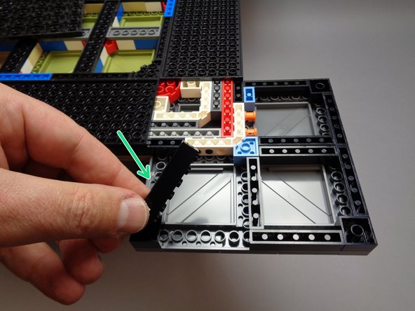 As shown by the green arrow in the first photo for this step, remove another 1x6 black brick, this time from the left side of the foot base.