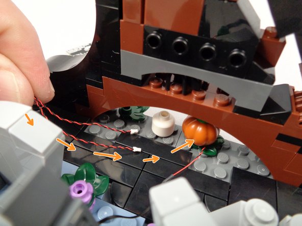 As shown by the orange arrows in the first photo for this step, pass the two pumpkin light wires under the gap in the wave base.