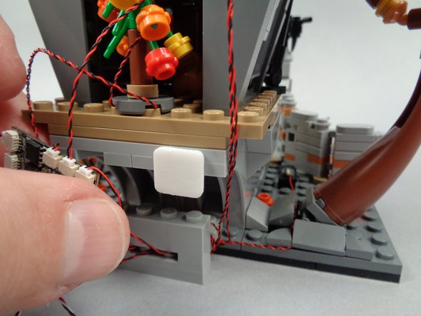 As shown in the photos for this step, take one of the large sticky squares included with your light kit and attach it to the back of Jack's house in the position shown in the second photo.