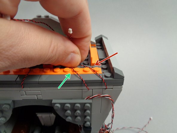 As shown by the red and green arrows in the first photo for this step, run the light wires for the banner light and right torch between studs on the long orange plate.