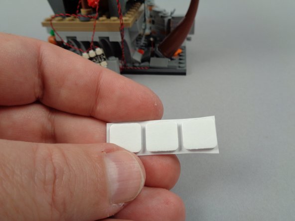 As shown in the photos for this step, take one of the large sticky squares included with your light kit and attach it to the back of Jack's house in the position shown in the second photo.