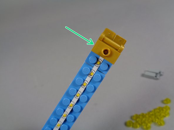 As shown by the green arrow in the first photo, take one of the 1x2 gold jumper plates included with your kit (the plates with holes drilled in the center), and use it to hold the light strip in place at the top of the marquee.