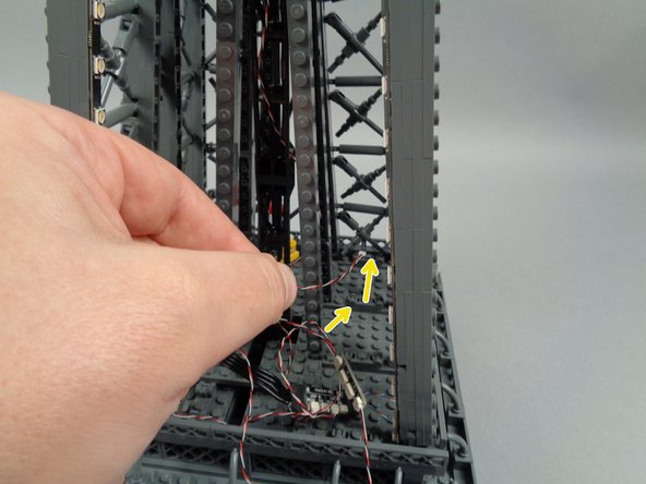 As shown by the yellow arrows in the second photo for this step, pass the other end of the control cable through the center of the tower toward the next pillar.