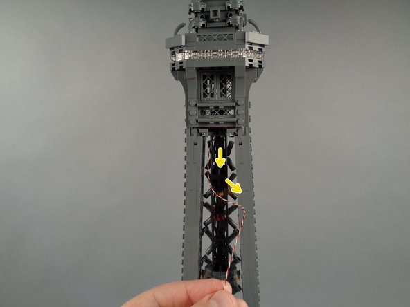 As shown by the yellow arrows in the photos for this step, carefully wind the control wire for the broadcast tower lights down through the center of the top tower section, looping the wire several times through the center black support pillars.