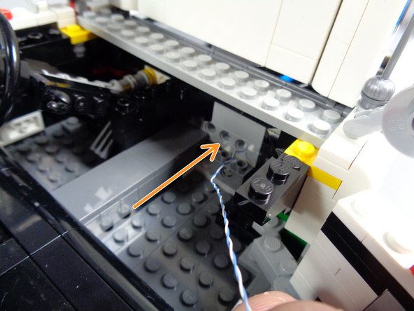 As shown by the orange arrow in the first photo for this step, pass the small plug for the speedometer light through one of the holes in the Technic brick behind the dashboard, on the side opposite the steering wheel.