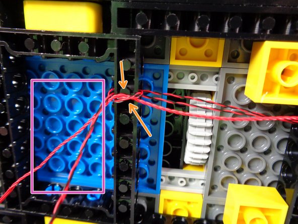 As shown by the green circle in the second photo for this step, pass the two strobe light wires through the holes in the black Technic frame behind the smaller steering gear.