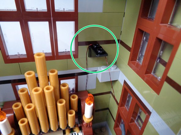 As shown by the green circle in the second photo for this step, run the wire for the first candle up from the organ, and loop it behind the back stud on the black slope piece.