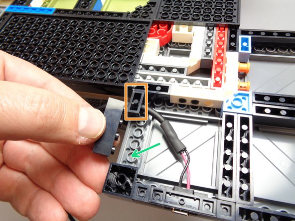 As shown by the orange rectangle in the first photo for this step, attach the 1x2 black brick with hole and power wire at the top of the base opening.