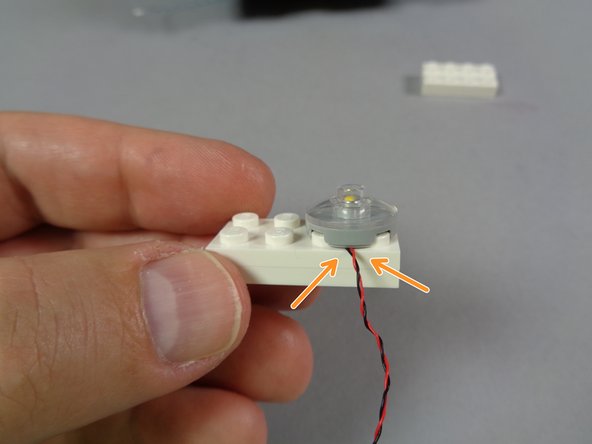 As shown in the second photo for this step, carefully re-attach the headlights to their white plate.  The orange arrows in the second photo show how the LED light wires should pass between, not on top of, studs.