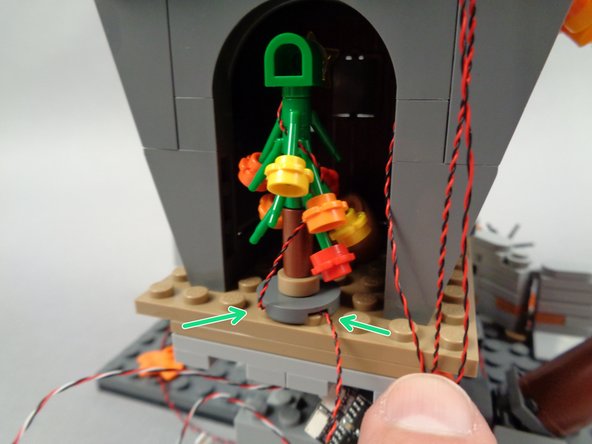 As shown by the green arrows in the third photo for this step, route the light wire underneath the round tree base plate, and then re-attach the tree to the floor of Jack's house.