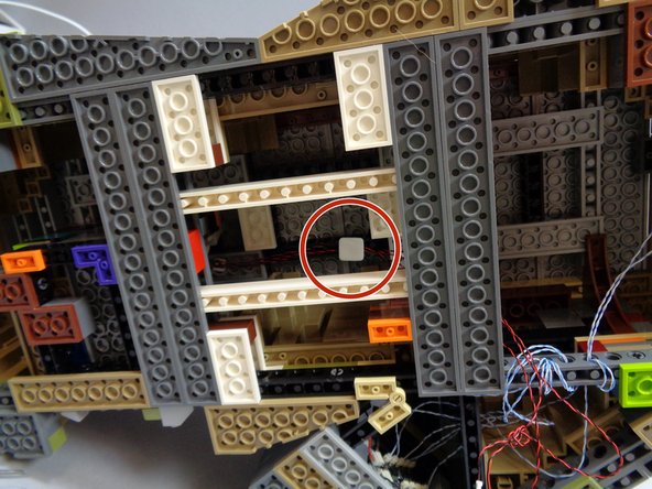 As shown by the red circles in the second and third photos, use one of the large sticky squares from Bag 1 to hold the three light wires to the top of the open space under the castle.