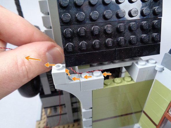 As shown by the orange arrows in the second photo for this step, pass the wire back along the edge of the wall and behind the 1x1 Technic brick on the end of the wall.