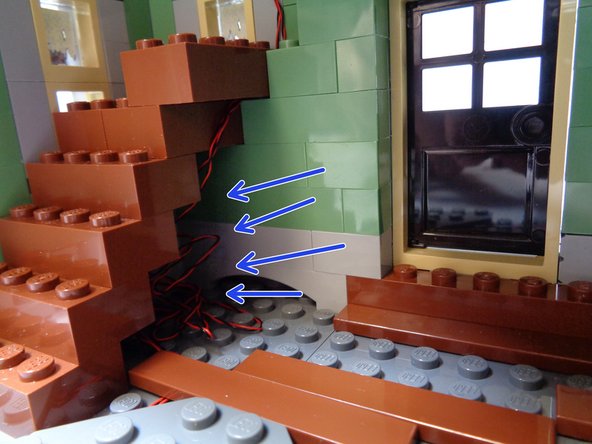 As shown in the second photo, re-attach the lowest 2x4 LEGO® brick on the staircase.  The brick should hold the two fireplace light wires in place along the front wall as shown by the orange arrow.