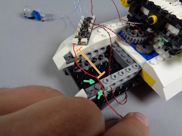 As shown by the green arrows in the second photo for this step, lay the red wire between the studs of the Technic brick and bracket.