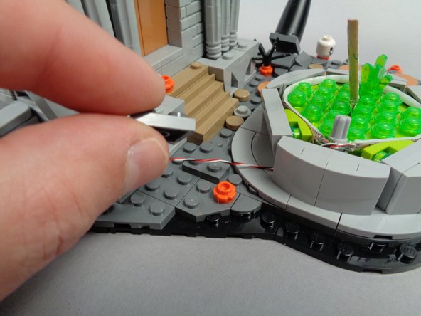 As shown in the first photo for this step, remove one of the dark bluish gray angle plates from the base behind the fountain, and pass the fountain light wire between the studs on the base.