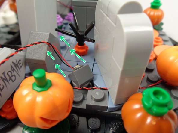As shown in the first photo, attach the second yellow LED light near the HENBANE tombstone and the rear lighted pumpkin, with the light facing back and on an angle as shown by the green arrows.