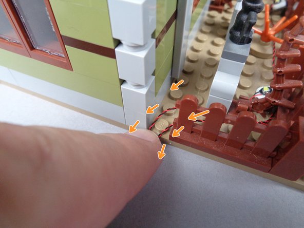 As shown by the two orange arrows in the second photo for this step, run the wires for the two lightning lights on either side of the baseplate stud under the fence post.