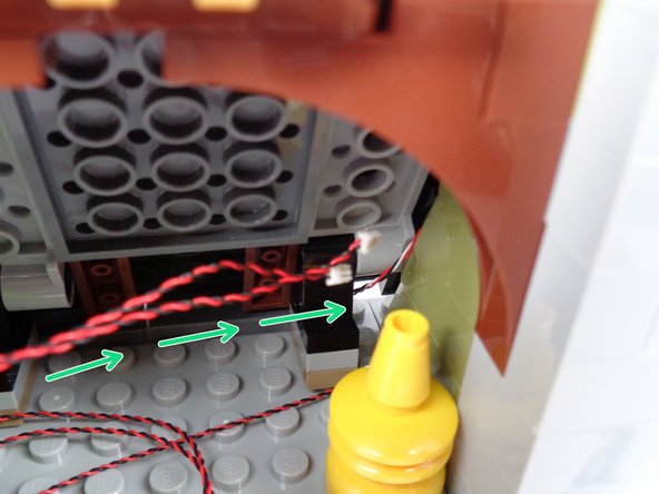 As shown by the green arrows in the third photo for this step, pass both red LED light wires through the gap between the roof and the back wall.