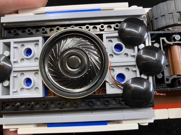 As shown in the second photo, press the speaker and sticky square down onto the &quot;boat stud&quot; you used in Step 4 to hold the wire coming from the front hood assembly.