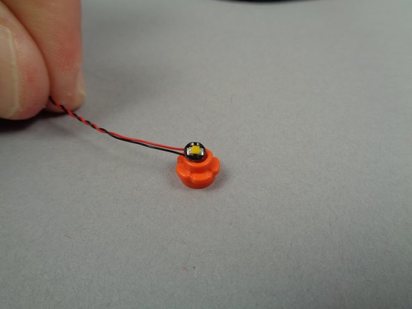 As shown in the third photo for this step, take one of the two remaining LED lights from the bag with the orange dot and center it on the flower plate with its light element facing upward.