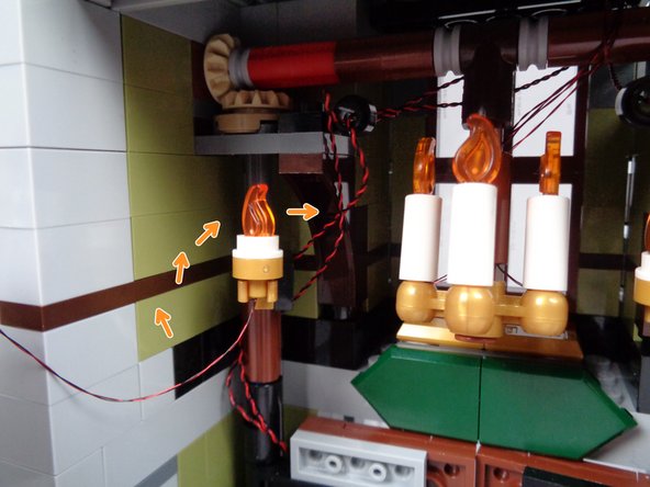As shown by the orange arrows in the first photo for this step, attach one of the pre-lit Brickstuff sconces in the gold LEGO clip to the left of the front door, and then loop the light wire once around the back of the door hinge as you did earlier for the fireplace and front porch lights.