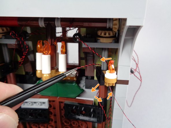 As shown by the orange arrows in the first photo for this step, attach the second pre-lit Brickstuff sconce in the gold LEGO clip to the right of the front door, and then loop the light wire once around the back of the door hinge as you did earlier for the front porch lights.