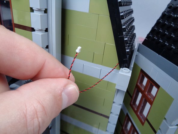 As shown by the blue arrows in the first photo for this step, pass the wire toward the back of the attic.  The plug should be able to fit through the small gap between the roof section and the green walls.