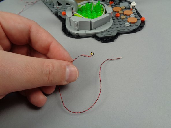 As shown in the first photo for this step, take one of the two remaining LED lights from the small pink bag with orange dot, and attach a small sticky dot to the back of the LED light as shown in the second photo.