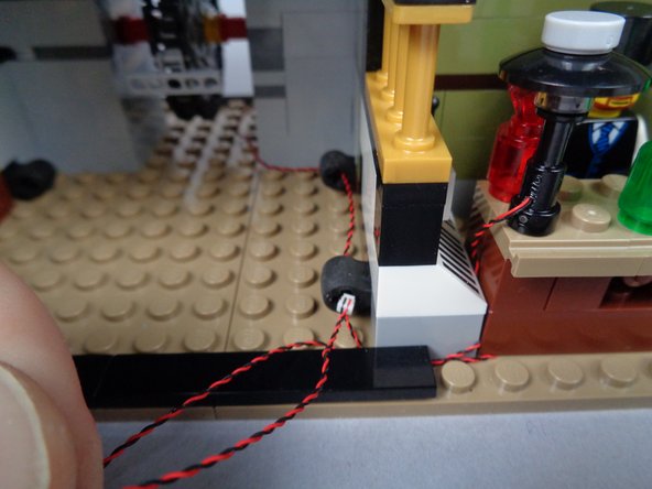 As shown in the third photo for this step, pass the lamp LED light wire back through the rubber Technic parts following the same path as the fireplace light wire.