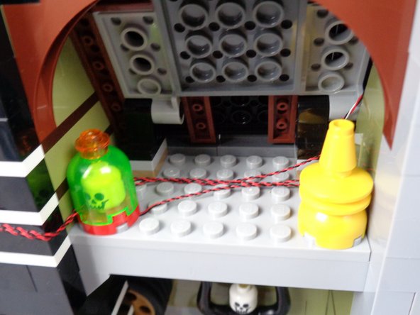 As shown in the first and second photos for this step, route the two red LED light wires behind the skull jar and toward the back of the attic.