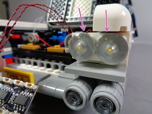 As shown in the first photo for this step, re-attach the first two headlights to the Ecto-1 body.  Note the two purple arrows in the photo, showing the light wires passing over the TOP of the white plate.