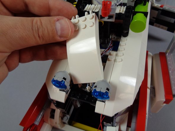 As shown in the second photo for this step, carefully and slowly slip the white roof slope between the two blue roof lights, working at an angle, until the white slope is centered and can be put back into place.