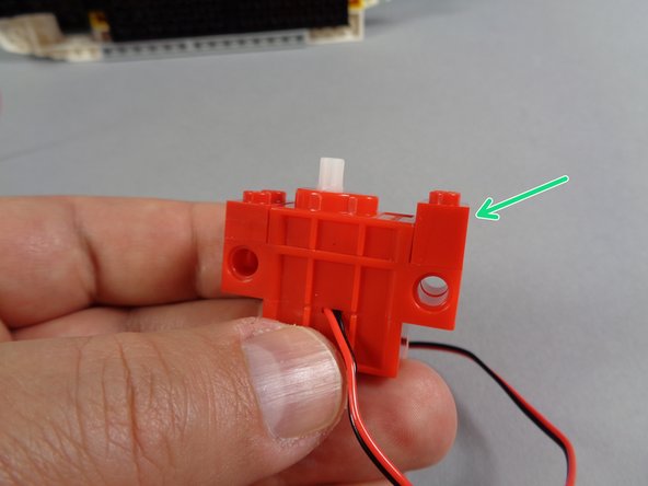As shown by the green arrow in the second photo for this step, attach one of the red 1x2 LEGO bricks to the motor on the opposite end as the plates.
