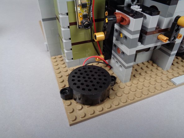 As shown in the first photo for this step, you can press the speaker down on top of the two black round 1x1 LEGO plates you mounted in the previous step.