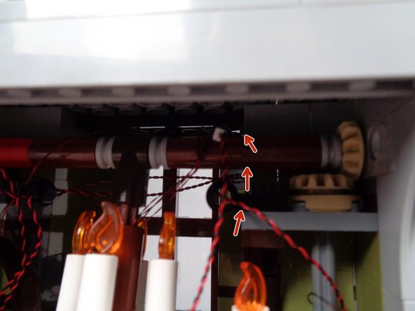 As shown by the red arrows in the second photo for this step, pass the plug for the light wire ABOVE and OVER the long axle that moves the front door.