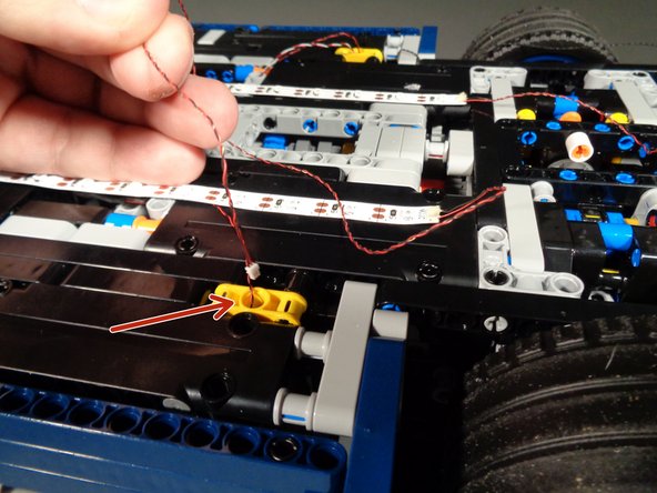 As shown by the red arrows in the first and second photos for this step, carefully feed the wires for both light strips through the hole in the center of the yellow Technic bracket on each side of the underframe.