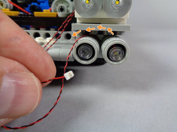 As shown by the orange arrows in the first photo for this step, re-attach the first lower headlight so the light wire passes out the top and toward the center of the Ecto-1.
