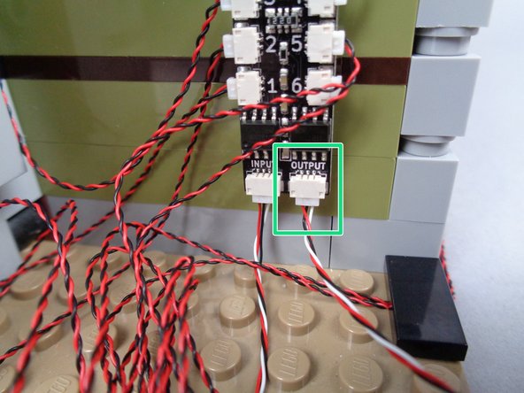 As shown by the green arrow and rectangle in the photos for this step, connect one end of the 3-wire connecting cable to the OUTPUT plug on the second BRANCH06X adapter board (the board you recently finished connecting lights to).