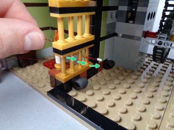 As shown by the green arrows in the second and third photos for this step, pass the light wire for the left candle through the space between the gold bars of the elevator shaft.