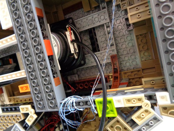 As shown by the red arrows in the third photo, carefully feed the speaker cable around and through the base of the castle, so the speaker cable passes through the same gap in the castle base as the 3-wire red connecting cable you ran in Step 7.