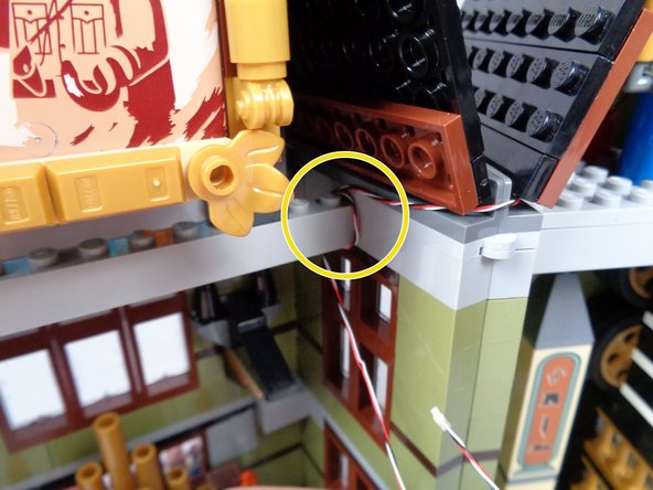 As shown by the green arrows in the second photo for this step, with the mansion opened up, route the 3-wire cable around to the front half of the mansion and loop the wire under the long light bluish gray brick.