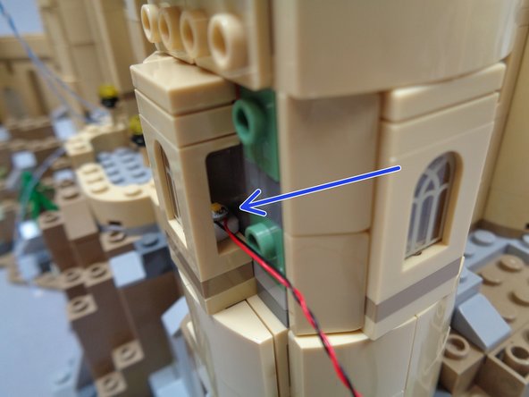 As shown in the third photo, take one of the warm white lights from Bag 10, attach a sticky square to the back, and fit it behind the lower front window as shown by the blue arrow in the third photo.