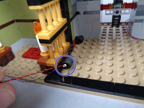 As shown in the photos for this step, carefully pass the plug for the first candle wire through the center of the two rubber Technic parts, toward the back of the elevator shaft.