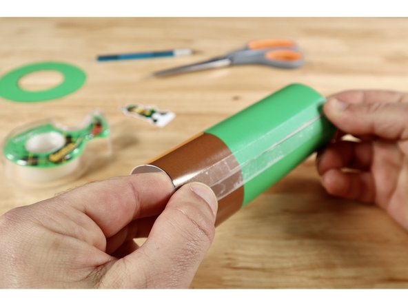 Using the brown belt as a reference for the bottom of the hat piece, curl it into a tube shape and attach at the seam with clear tape. You may benefit from pulling the flat sheet against the edge of a table to begin coaxing it into the tube shape before taping.