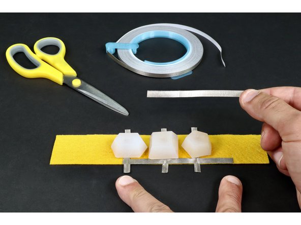 Now, cut a piece of Maker Tape that can reach across/atop all three LED legs on a single side plus about 1/2&quot; beyond . Peel and stick it atop/across the shorter negative legs as pictured in photo 1.