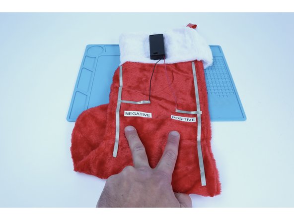 Stick one end of one to any spot along the length of one of the longer side paths, then inward atop the stocking fabric, then doubled back on itself around the exposed wire of the nearest battery lead. Repeat this step with the remaining piece of Maker Tape for the opposite side.