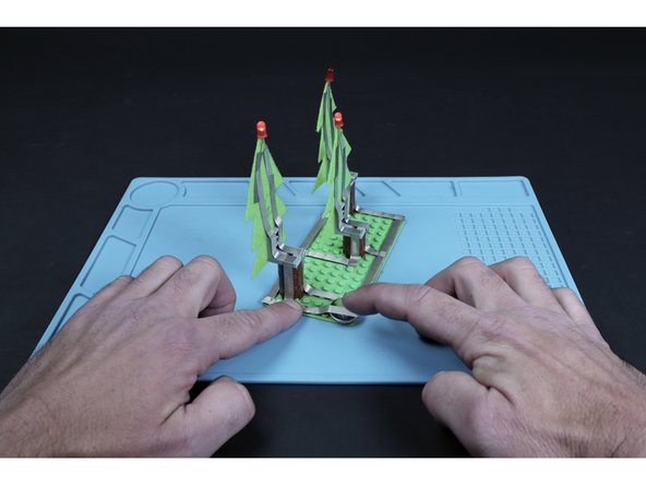 Measure, cut, peel and stick one final tape length where shown so that it can be stuck to the top of the battery. This will complete the circuit and turn on all three of your blinking LEDs. Peeling it away from the top of the battery will turn the circuit off.