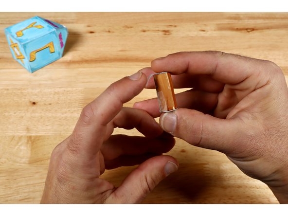 Use clear tape to finish and secure sides of cylindrical dreidel handle.  Start with the long seam that helps to turn the curly rectangle into a tube. Then fold the end caps into place and secure them with clear tape as shown.