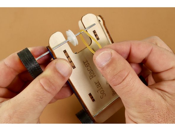 Find and set aside the large plastic axle wheel, rubber band, and final wheel. Thread the remaining wheel axle/wheel combo through the FIRST drop-down hole of the remaining pair.  Before threading this axle rod through the final drop-down hole, thread it through both the plastic wheel AND rubber band.