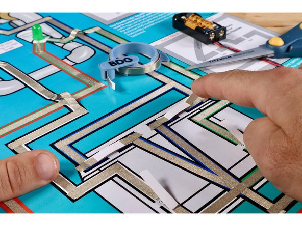 Install the switches by positioning them atop the gaps for the Blue LED/ROV outcome circuit (Maker Tape side down) while securing each with another 2 pieces of Maker Tape as shown. The LED should now momentarily light up when a given pushbutton is pressed.